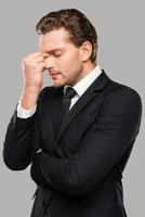 Too stressful day. Frustrated young man in formalwear touching head with fingers and keeping eyes closed while standing against grey background photo