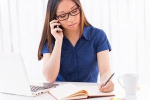 Busy working. Beautiful young Asian woman writing something in her note pad and talking on the mobile phone while sitting at her working place photo