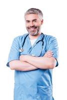 Experienced and confident surgeon. Cheerful mature doctor looking at camera and smiling while keeping arms crossed and standing isolated on white photo