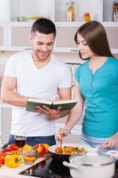 aprendiendo a cocinar juntos. feliz pareja joven cocinando juntos en la cocina mientras el hombre sostiene un libro de cocina foto