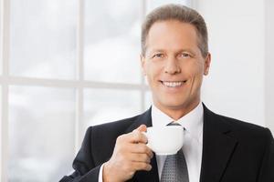 Coffee break. Portrait of cheerful mature man in formalwear holding coffee cup and smiling to you while standing near window photo