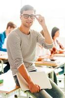 estudiante confiado. joven sonriente sosteniendo una laptop y ajustándose los anteojos mientras se inclina en el escritorio foto