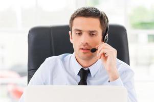 Operator at work. Confident young man looking at laptop and adjusting his headset while sitting at his working place photo