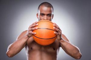 el baloncesto es mi vida. joven africano sin camisa escondiendo parte de su rostro detrás de la pelota de baloncesto y mirando a la cámara mientras se enfrenta a un fondo gris foto