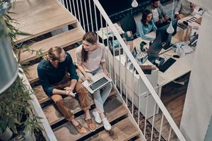 Top view of two young modern colleagues in smart casual wear communicating and using laptop while sitting on the stairs photo