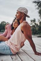 amar por siempre. feliz pareja joven abrazándose y sonriendo mientras se sienta en el muelle cerca del lago foto