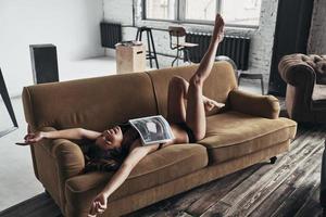 Lazy morning. Attractive young woman covered with magazine smiling and gesturing while lying on the sofa at home photo
