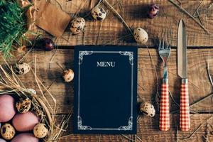 Easter dinner menu. Top view of Easter eggs and menu board with fork and knife lying on wooden rustic table with hay photo