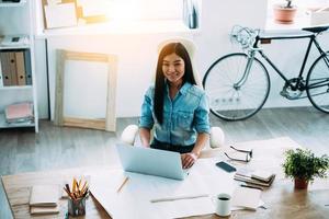 Working day of modern architect. Beautiful young Asian woman using laptop and looking at camera with smile while sitting at her working place photo