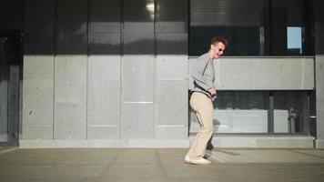 un apuesto hombre divertido y feliz con gafas de sol corre activamente, sonríe y baila con alegría y felicidad a lo largo de la calle de la ciudad. escena rodada en un parque urbano junto al centro de negocios. camara lenta video