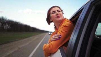 Fröhliches schönes Mädchen, das auf dem Rücksitz eines Autos fährt, schaut aus dem offenen Fenster und lächelt. Reisende Frau genießt die atemberaubende Landschaft. Zeitlupe. video
