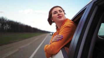 heureuse belle fille à cheval sur le siège arrière d'une voiture regarde par la fenêtre ouverte et sourit. femme voyageuse apprécie le paysage magnifique. ralenti. video