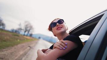 Fröhliches schönes Mädchen mit Sonnenbrille, das auf dem Rücksitz eines Autos fährt, schaut aus dem offenen Fenster und lächelt am Sommertag. reisende freudige frau genießt die atemberaubende landschaft an einem hellen sonnigen tag. Zeitlupe. video
