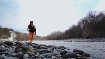 hermosa mujer de moda con gafas de sol camina por la orilla rocosa de un río de montaña. estilo de vida y viajes. camara lenta. video