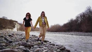 två lesbisk kvinnor håll händer och promenad längs en skön berg flod. två kärleksfull flickvänner. begrepp HBTQ, lycka, frihet, homosexuell par. långsam rörelse. video