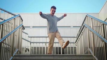 Handsome happy man in sunglasses is actively running, smiling and dancing with joy and happiness going down the city stairs. Scene Shot in an Urban Park Next to Business Center. Slow motion. video