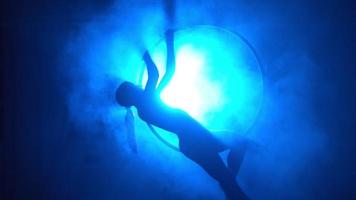 silueta, mujer gimnasta aérea realiza un truco en el ring en una habitación llena de humo con luz azul retroiluminada. iluminación de neón video