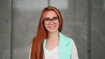 Portrait, beautiful young happy woman in glasses looks at camera and smiles. Female office worker with red hair in turquoise suit stands near business center. Student laughs in good mood. Slow motion. video