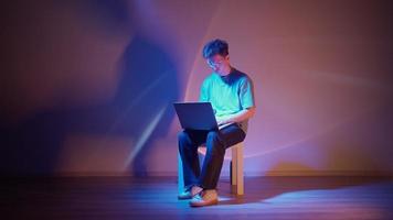 Lonely stylish man in glasses sits working at a laptop in neon lighting. The concept of modern technology and remote work as a freelancer.