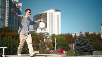 knap gelukkig Mens in zonnebril is actief rennen, glimlachen en dansen met vreugde en geluk langs de stad straat. tafereel schot in een stedelijk park De volgende naar bedrijf centrum. langzaam beweging. video