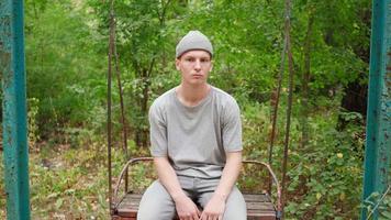 A sad guy sits on an abandoned swing in the forest and sways back and forth video