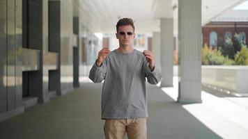 Handsome serious man takes off his black sunglasses and looks at the camera. Caucasian stylish young guy on the background of a city street. Casual style of clothing. Slow motion. video