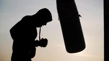 silhueta de um boxeador se exercitando ao ar livre. lutador atinge um saco de pancadas ao pôr do sol. forte homem atlético está envolvido em artes marciais na rua. fitness e conceito de estilo de vida saudável. video