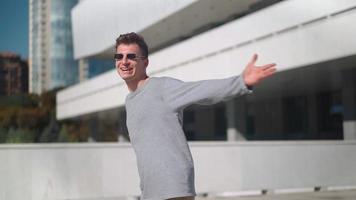 Happy handsome man in sunglasses raises his hands up and spins with happiness against background city. Caucasian guy smiling and laughing rejoicing in success and freedom in urban park. Slow motion. video