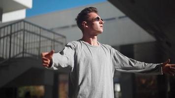 Handsome stylish white man in sunglasses raises his hands to the sides against the background of a city street. Tough confident Caucasian guy. Concept Motivation and freedom. Slow motion video