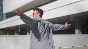 feliz hombre guapo con gafas de sol levanta las manos y gira con felicidad contra la ciudad de fondo. chico caucásico sonriendo y riendo regocijándose en el éxito y la libertad en el parque urbano. camara lenta. video