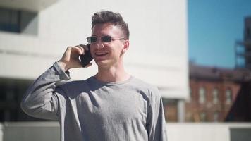 un apuesto hombre caucásico con gafas de sol camina y habla por teléfono móvil en un parque de la ciudad. joven feliz riendo y sonriendo mientras habla por teléfono al aire libre. camara lenta. video