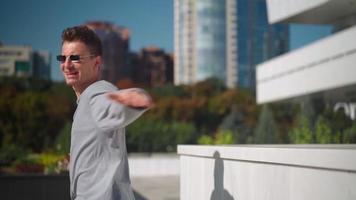 un apuesto hombre feliz con gafas de sol corre activamente, sonríe y baila con alegría y felicidad a lo largo de la calle de la ciudad. escena rodada en un parque urbano junto al centro de negocios. camara lenta. video