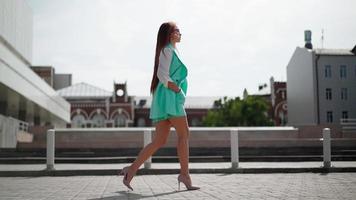 un' bellissimo sottile donna nel bicchieri e un' di moda turchese completo da uomo passeggiate attraverso centro. bella sexy attività commerciale signora con snello gambe nel alto tacchi è a piedi giù il città strada. lento movimento. video