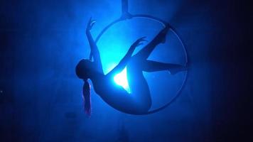 Silhouette, woman aerial gymnast performs a trick in the ring in a smoky room with backlit blue light. Neon lighting. video