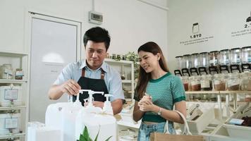 Servicemitarbeiter in einem Einzelhandelsgeschäft, asiatischer männlicher Ladenbesitzer, der wiederverwendbare Behälter drückt, um flüssige Produkte an einen Kunden im Nachfüllladen zu füllen, abfallfreie Lebensmittel, Bio-Produkte, nachhaltiger Lebensstil. video