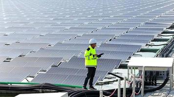 electricista masculino con casco de seguridad blanco parado en una escalera y montando un panel solar fotovoltaico bajo un hermoso cielo azul. concepto de fuentes alternativas de energía. video