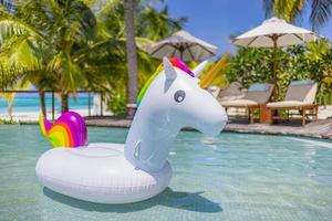 Inflatable colorful white unicorn at resort swimming pool. Vacation time in tropical pool with plastic toys. Relaxation and fun concept, panoramic summer background. Palm trees, chairs, beds umbrellas photo