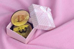 The golden bitcoin lies in a small pink gift box with a small bow on a blanket made of soft and fluffy light pink fleece fabric with a large number of relief folds photo