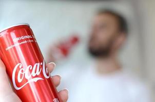 KHARKOV. UKRAINE - MAY 2, 2019 Caucasian man drinks Coca-Cola drink in garage interior and male hand presents one Coca cola red can in focus photo