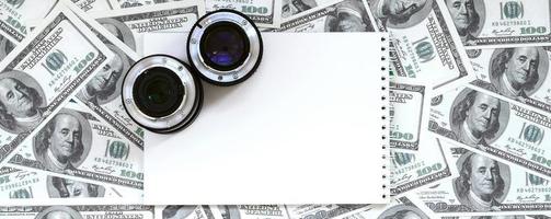 dos lentes fotográficos y un cuaderno blanco se encuentran en el fondo de muchos billetes de dólar. espacio para texto foto