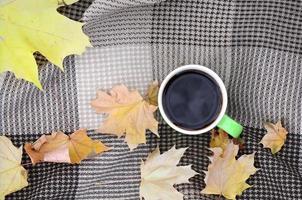 hojas de otoño y una taza de café caliente y humeante se encuentran en cuadros escoceses al aire libre foto