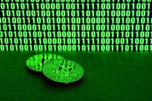 A pair of bitcoins lies on a cardboard surface on the background of a monitor depicting a binary code of bright green zeros and one units on a black background. Low key lighting photo