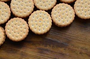 A round sandwich cookie with coconut filling lies in large quantities on a brown wooden surface. Photo of edible treats on a wooden background with copy space