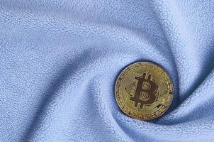 The golden bitcoin lies on a blanket made of soft and fluffy light blue fleece fabric with a large number of relief folds. The shape of the folds resembles a fan from a video card cooler photo