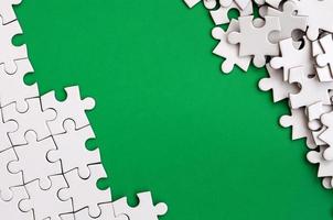 Fragment of a folded white jigsaw puzzle and a pile of uncombed puzzle elements against the background of a green surface. Texture photo with space for text