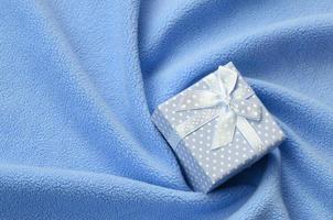 A small gift box in blue with a small bow lies on a blanket of soft and furry light blue fleece fabric with a lot of relief folds. Packing for a gift to your lovely girlfriend photo