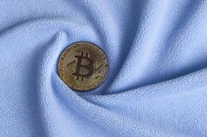 The golden bitcoin lies on a blanket made of soft and fluffy light blue fleece fabric with a large number of relief folds. The shape of the folds resembles a fan from a video card cooler photo