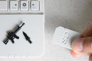 A hand inserts a compact SD card into the slot of a netbook, on which lie a miniature knife and gun. The concept of information warfare, journalistic activity, dissemination of provocative information photo