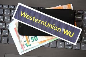 TERNOPIL, UKRAINE - SEPTEMBER 6, 2022 Western Union paper logotype lies on black laptop with euro bills. Western Union Company is American multinational financial services company photo