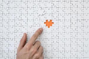 The texture of a white jigsaw puzzle in the assembled state with one missing element, forming an orange space, pointed to by the finger of the male hand photo